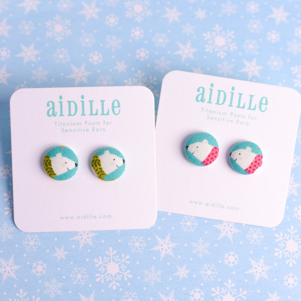 Pair of earrings with polar bear design on cards with 'aidille' branding against a snowflake patterned background.