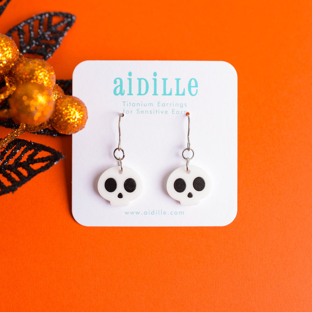 Skull-shaped earrings on a 'aidille' branded card against an orange background