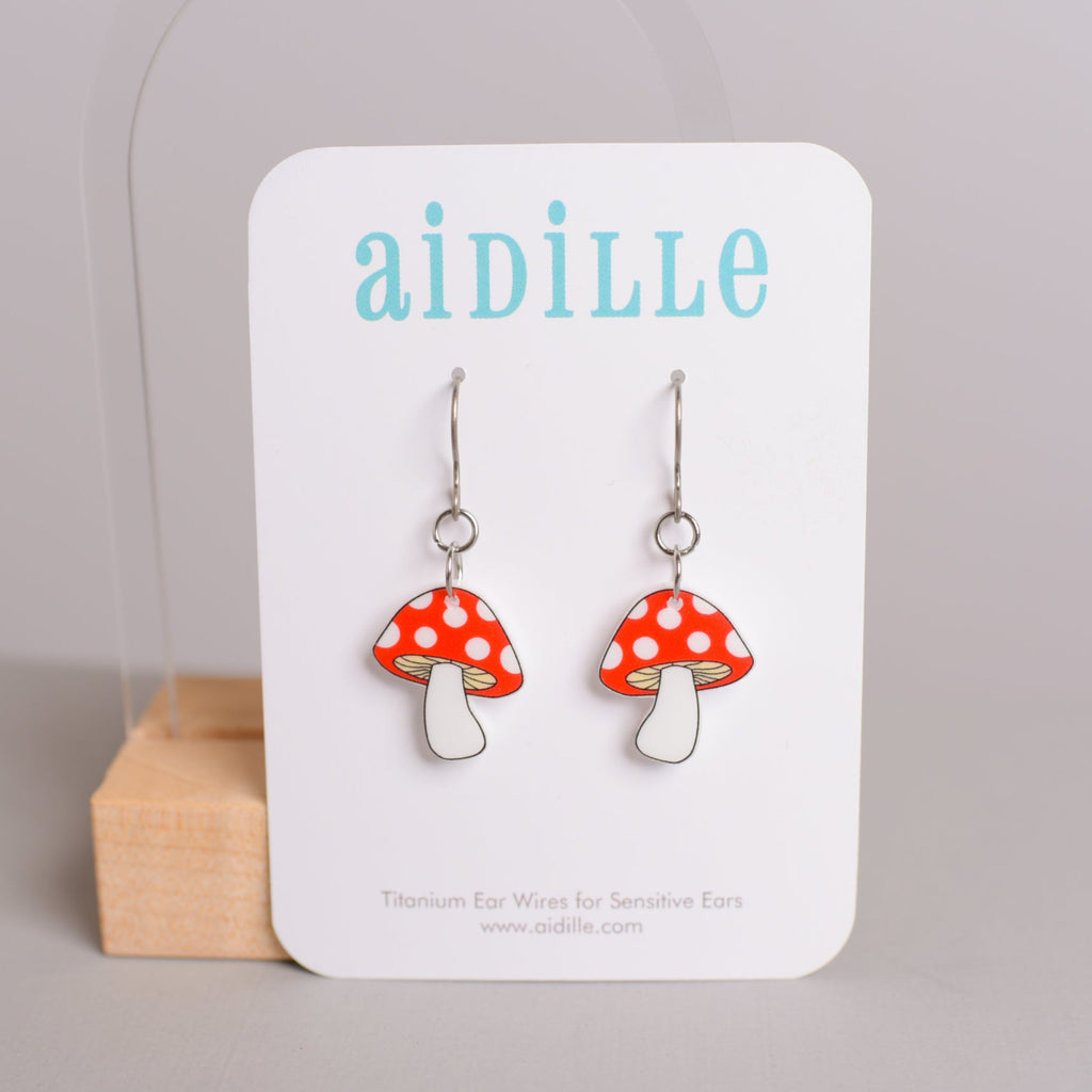 Mushroom-shaped earrings on a 'aidille' branded card with a wooden block in the background.