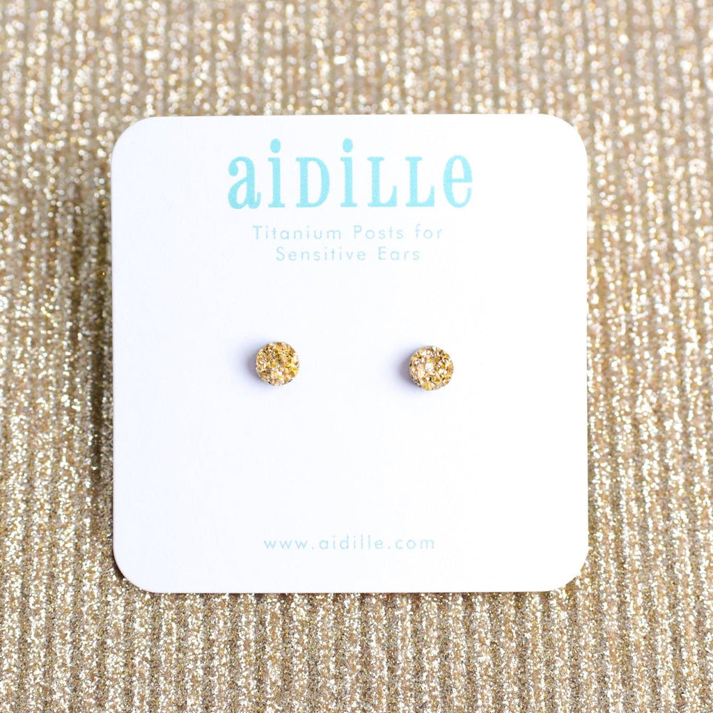 Gold stud earrings on a 'aidille' branded card against a glittery beige background