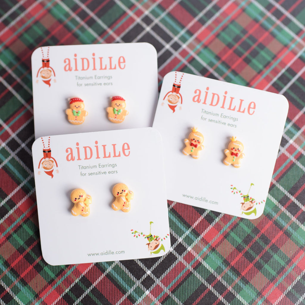 Three pairs of gingerbread man earrings on 'aidille' branded cards against a plaid background.