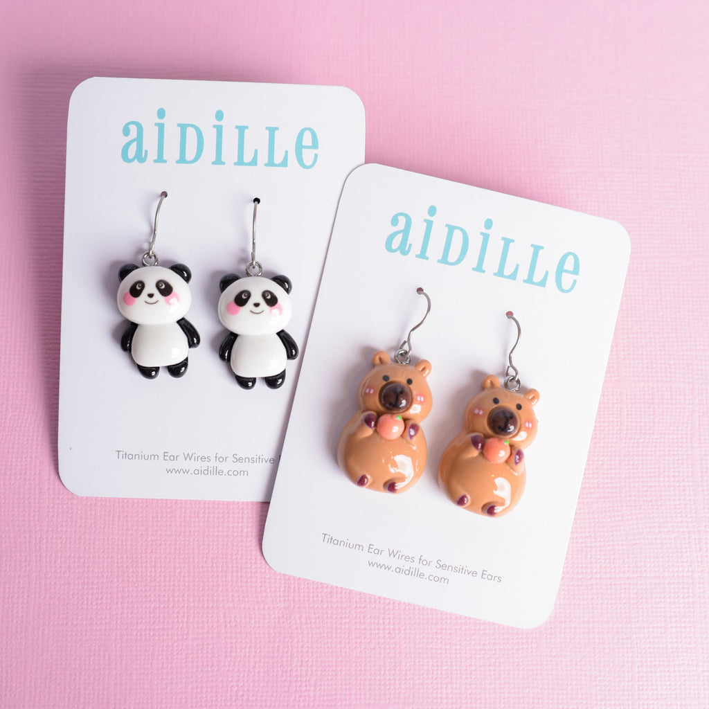 Panda bear and capybara earrings on 'aidille' branded cards against a pink background