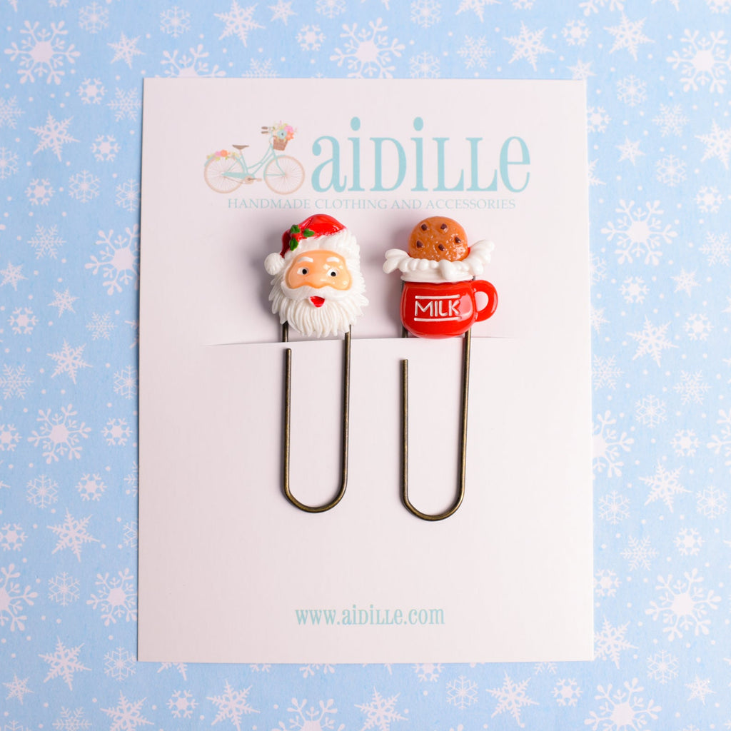 Two paper clips shaped like Santa Claus and a milk cup on a card with 'aidille' branding against a snowflake-patterned background.