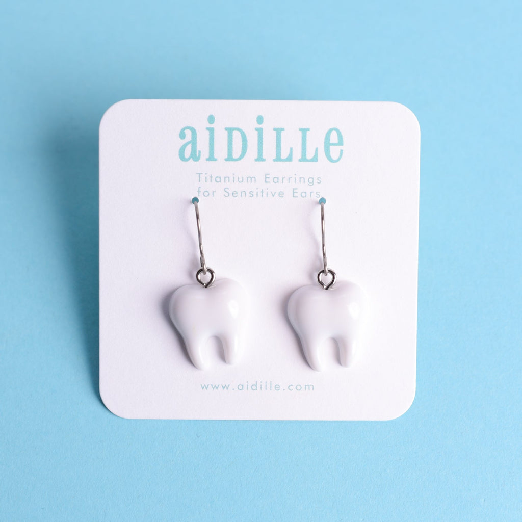 Pair of tooth-shaped earrings on a 'aidille' branded card against a light blue background