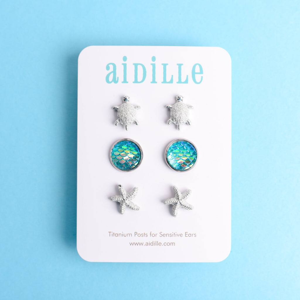 Set of turtle, starfish, and blue opal earrings on a 'aidille' branded card against a light blue background.