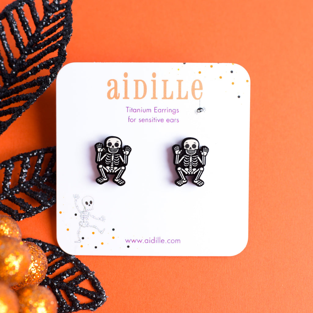 Skeleton earrings on a 'aidille' branded card with Halloween decorations on an orange background