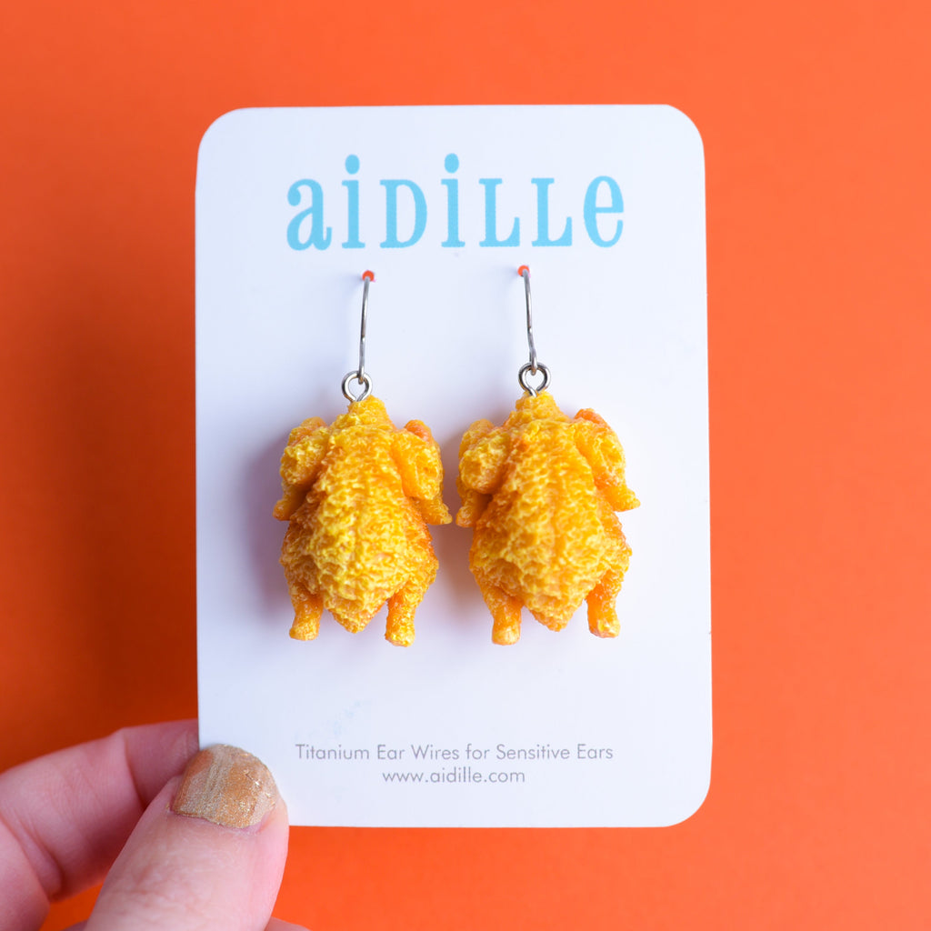 Yellow turkey-shaped earrings on a 'aidille' branded card against an orange background