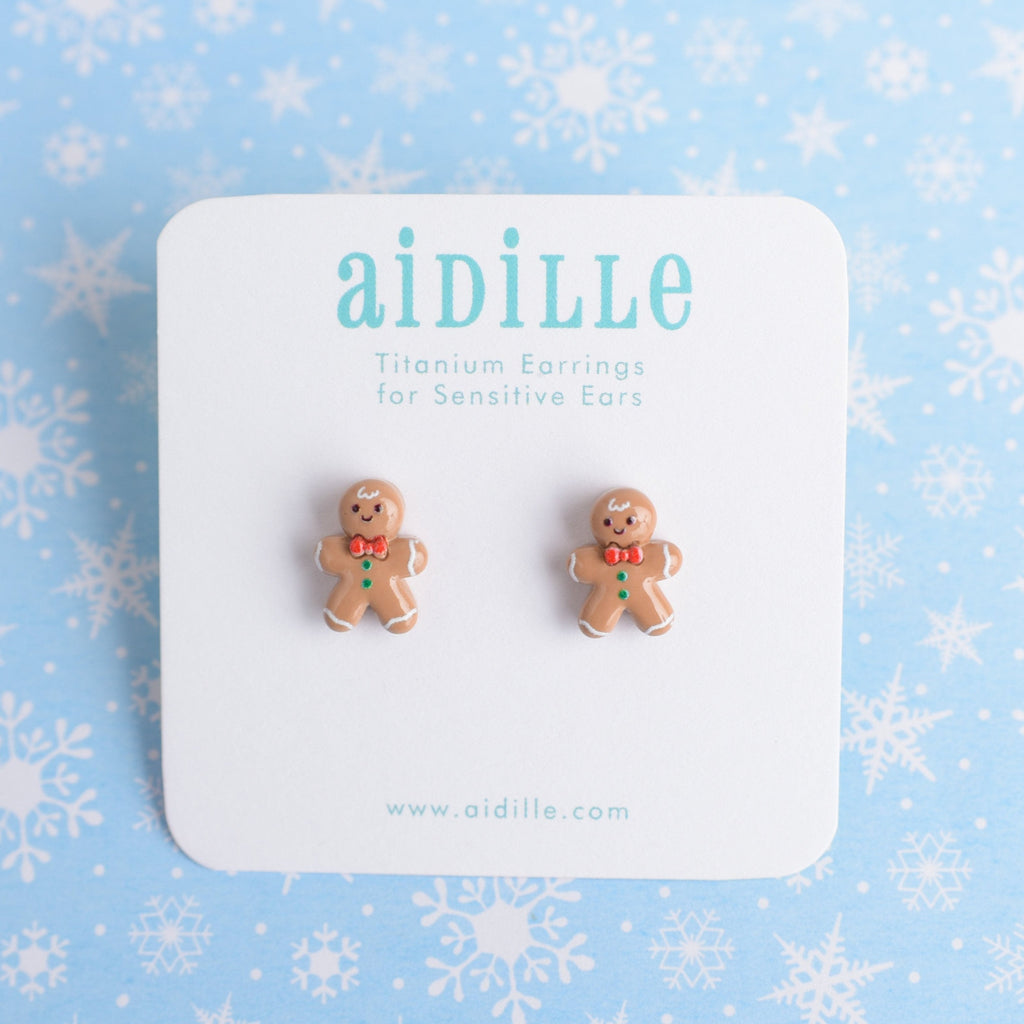 A pair of chunky resin gingerbread man earrings with hypoallergenic titanium posts, displayed on a white card with snowflake pattern in the background.